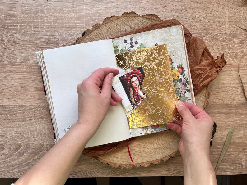 May include: A person is holding a gold leaf sheet over a page in a journal. The page has a floral design and a picture of a woman wearing a red and gold crown.