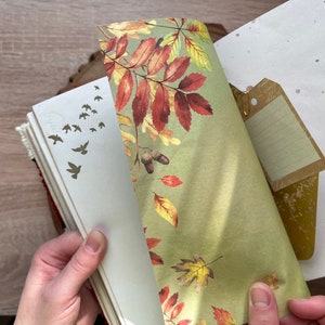 May include: A close-up of a journal with a green and brown patterned paper bookmark. The journal has a white cover with gold leaf accents and a small tag attached to the top.