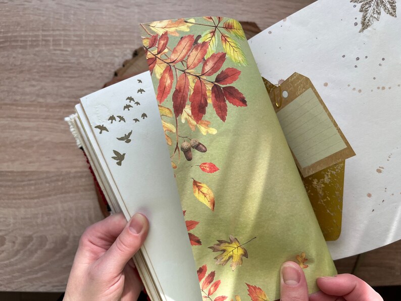 May include: A close-up of a journal with a green and brown patterned paper bookmark. The journal has a white cover with gold leaf accents and a small tag attached to the top.