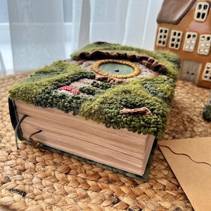 Op de afbeelding: Een boek met een groene, bruine en gele kaft met een afbeelding van een ronde opening met een groene achtergrond. Het boek is dicht en ligt op een geweven strooien mat.