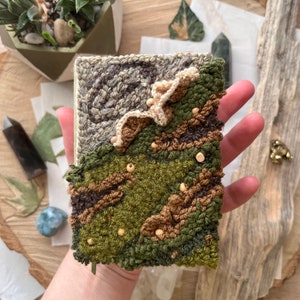 May include: A small, hand-stitched wall hanging with a green, brown, and beige color scheme. The piece features a textured, abstract design with a raised, textured surface.