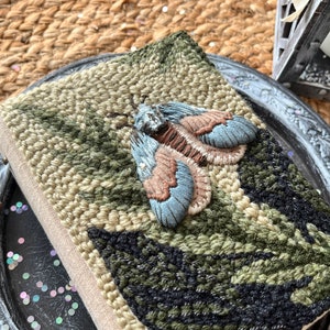 May include: A hand-stitched book cover featuring a detailed embroidered moth design in shades of blue, green, and brown. The cover is made from natural linen fabric and has a textured surface.