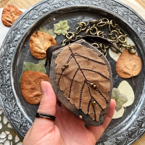 Op de afbeelding: Een bruine stoffen tas in de vorm van een blad met een ketting eraan. De tas heeft een gestikt ontwerp en is versierd met kleine gouden kralen.