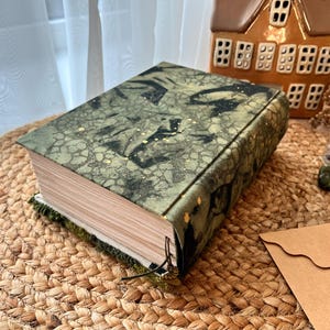 K&ouml;nnte beinhalten: Ein gr&uuml;nes und goldenes Buch mit einem strukturierten Einband und einem schwarzen Lederriemen. Das Buch ist geschlossen und liegt auf einer gewebten Matte.
