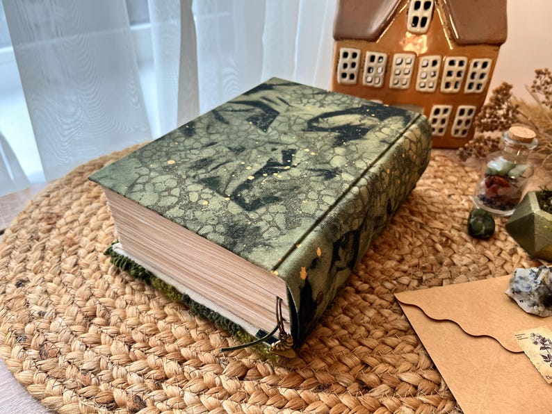 K&ouml;nnte beinhalten: Ein gr&uuml;nes und goldenes Buch mit einem strukturierten Einband und einem schwarzen Lederriemen. Das Buch ist geschlossen und liegt auf einer gewebten Matte.