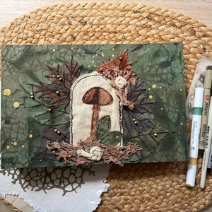 May include: A handmade journal with a forest theme. The cover is green with a mushroom and leaf design, embellished with brown and gold accents. Art supplies, including paintbrushes and pens, are arranged nearby on a woven mat.