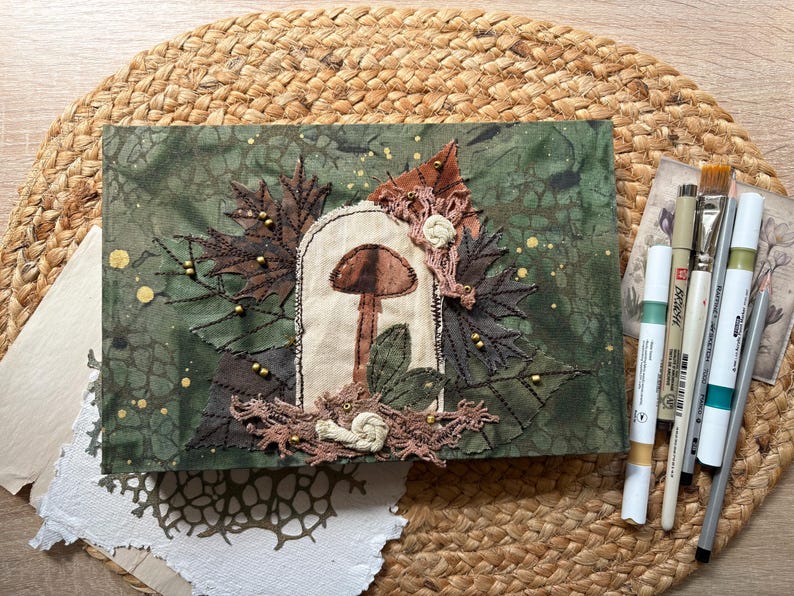 May include: A handmade journal with a forest theme. The cover is green with a mushroom and leaf design, embellished with brown and gold accents. Art supplies, including paintbrushes and pens, are arranged nearby on a woven mat.
