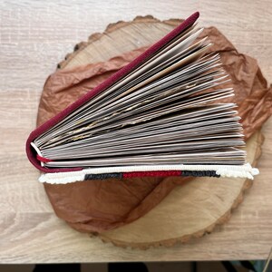 May include: A burgundy leather-bound journal with a white, black and red embroidered trim. The journal is open and fanned out, revealing blank pages.