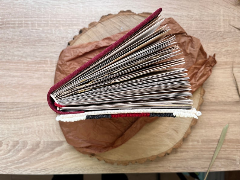 May include: A burgundy leather-bound journal with a white, black and red embroidered trim. The journal is open and fanned out, revealing blank pages.