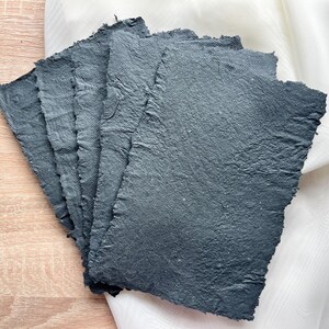 May include: A stack of six sheets of handmade, dark gray paper with deckled edges. The paper has a rough, textured surface.