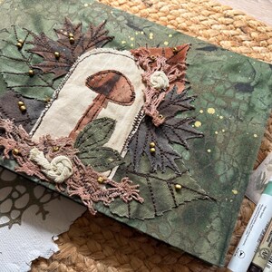 May include: A handmade journal with a green and gold speckled cover. The cover features an embroidered mushroom design with brown and beige accents, surrounded by fabric leaves and lace. Decorative beads add detail.