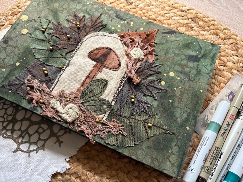 May include: A handmade journal with a green and gold speckled cover. The cover features an embroidered mushroom design with brown and beige accents, surrounded by fabric leaves and lace. Decorative beads add detail.