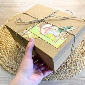 K&ouml;nnte beinhalten: Ein brauner Karton mit einem dekorativen Papierdesign mit Pilzen und Bl&auml;ttern. Die Schachtel ist mit Schnur gebunden und steht auf einer gewebten Matte.