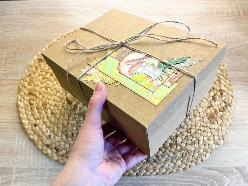 K&ouml;nnte beinhalten: Ein brauner Karton mit einem dekorativen Papierdesign mit Pilzen und Bl&auml;ttern. Die Schachtel ist mit Schnur gebunden und steht auf einer gewebten Matte.