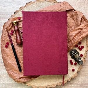 May include: A burgundy velvet journal with a red ribbon bookmark, a wooden stamp with a brass handle, a black ink pen, and a metal seal press with a hand design. The items are arranged on a wooden slice with brown paper and red wax seals.