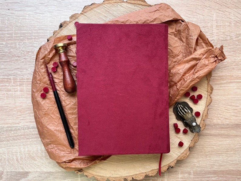May include: A burgundy velvet journal with a red ribbon bookmark, a wooden stamp with a brass handle, a black ink pen, and a metal seal press with a hand design. The items are arranged on a wooden slice with brown paper and red wax seals.