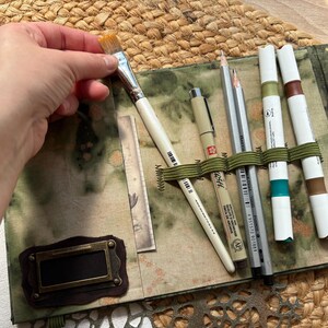 May include: An open art supply case with a variety of tools. A hand holds a paintbrush over the case. The case contains pens, pencils, and markers secured by an olive green elastic band. The case has a green and brown patterned fabric exterior.