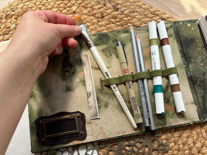 May include: An open art supply case with a variety of tools. A hand holds a paintbrush over the case. The case contains pens, pencils, and markers secured by an olive green elastic band. The case has a green and brown patterned fabric exterior.