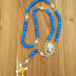May include: A blue beaded rosary with a gold cross and decorative centerpiece. The beads are a vibrant blue, with white and gold accents. The cross and centerpiece feature detailed religious imagery.
