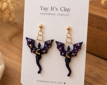 May include: A pair of dark purple dragon-shaped earrings with gold foil accents, suspended from gold-toned studs on a white display card labeled Yay It's Clay Handmade Jewelry. The earrings feature detailed wings and tails with a celestial pattern.