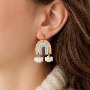 Könnte beinhalten: Ein Hängeohrring mit einem pastellfarbenen Regenbogenbogen in Pink, Gelb und Hellblau, der an einem goldfarbenen Stecker befestigt ist. Zwei kleine, weiße, wolkenförmige Anhänger hängen an der Basis des Regenbogens, verbunden durch goldfarbene Metallringe.
