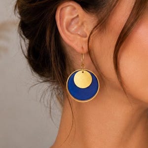 Könnte beinhalten: Goldfarbene Creolen mit blauem und goldenem Kreisdesign. Die Ohrringe haben einen großen blauen Kreis mit einem kleineren goldenen Kreis darüber. Ein modernes und stilvolles Accessoire.