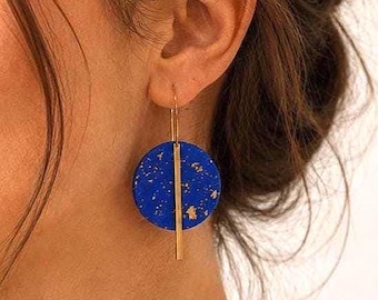 May include: A close-up of a blue and gold earring. The earring has a circular design with gold flecks and a vertical gold bar. The earring is hanging from a gold hoop.