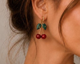 May include: Gold hoop earrings with a cherry design. Each earring features two red cherries and green leaves. The earrings are a fun and playful accessory.