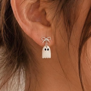 Può includere: Un orecchino color argento caratterizzato da un piccolo ciondolo a forma di fantasma bianco con due occhi neri, sospeso a un delicato perno a forma di fiocco color argento. Il fantasma ha una parte superiore arrotondata e un bordo inferiore.