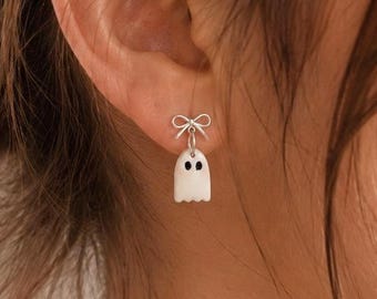 May include: A silver-toned earring featuring a small, white ghost charm with two black eyes, suspended from a delicate silver-toned bow stud. The ghost has a rounded top and a scalloped bottom edge, creating a simple, clean design for the ear.