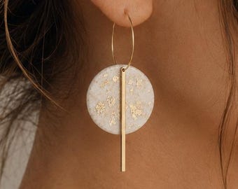 May include: A dangle earring featuring a thin gold hoop attached to a circular white disc with gold leaf flecks. A vertical gold bar hangs from the center of the disc, extending downward. The earring is suspended from a gold wire hook.
