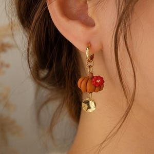 Könnte beinhalten: Ein goldener Creolen-Ohrring mit einem kleinen, orangefarbenen, gerippten Kürbis-Anhänger, der mit einer winzigen roten Blume verziert ist, sowie einer kleinen, runden, gehämmerten goldfarbenen Metallscheibe, die unter dem Kürbis hängt.