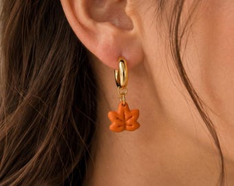 May include: A gold hoop earring featuring a small, matte orange maple leaf charm. The charm is suspended from a polished gold-tone circular hoop that secures through the earlobe. The leaf design includes subtle embossed vein details on its surface.