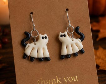 May include: A pair of dangle earrings featuring white cat-shaped charms with black tails and paws, suspended from silver-toned metal hooks. The earrings are attached to a rectangular brown card with the text thank you and a small heart icon.