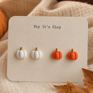 Può includere: Due paia di orecchini a lobo a forma di zucca fissati su un cartoncino rettangolare con la scritta Yay It's Clay. Un paio è bianco e l'altro è arancione, entrambi con piccoli steli color oro sulla parte superiore delle zucche.
