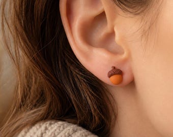 May include: A small stud earring shaped like an acorn, featuring a textured dark brown cap and a smooth, rounded light orange nut. The earring is secured to the earlobe, showcasing a detailed, miniature representation of a natural acorn.
