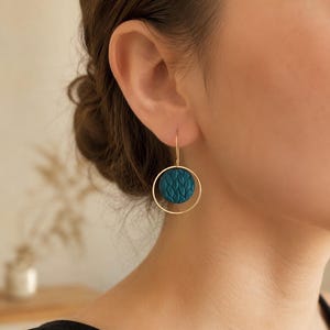 May include: A gold-toned earring featuring a thin circular hoop frame that surrounds a smaller, circular teal-colored disc. The teal disc has a raised, repeating leaf-like texture across its surface. The earring hangs from a simple gold wire hook.