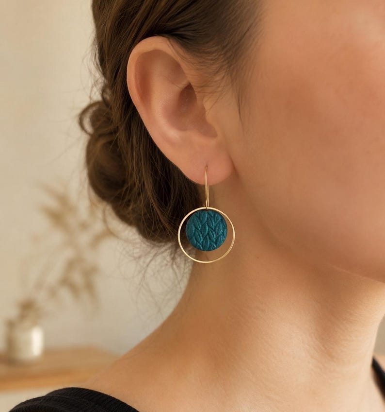 May include: A gold-toned earring featuring a thin circular hoop frame that surrounds a smaller, circular teal-colored disc. The teal disc has a raised, repeating leaf-like texture across its surface. The earring hangs from a simple gold wire hook.