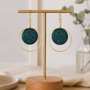 May include: A pair of dangle earrings featuring dark teal circular discs with a textured leaf pattern suspended within thin gold-toned metal hoops. The earrings hang from gold-toned hooks and are displayed on a gold metal T-bar stand.