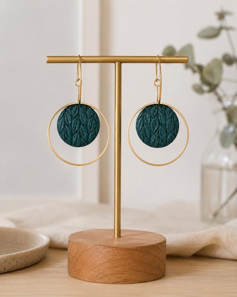May include: A pair of dangle earrings featuring dark teal circular discs with a textured leaf pattern suspended within thin gold-toned metal hoops. The earrings hang from gold-toned hooks and are displayed on a gold metal T-bar stand.