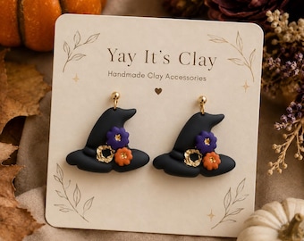 May include: A pair of black witch hat earrings made of clay, featuring small purple and orange flowers and gold-colored accents. The earrings are displayed on a cream-colored card with the text Yay It's Clay Handmade Clay Accessories.