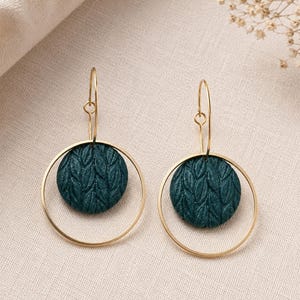 May include: A pair of dangle earrings featuring a circular gold-toned wire frame and a central dark teal circular disc. The disc has a textured surface with a repeating leaf-like pattern. The earrings hang from thin gold-toned curved ear wires.