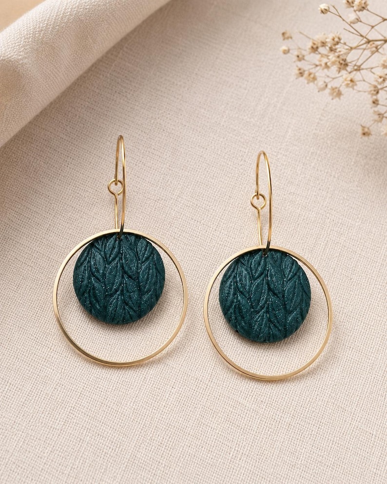May include: A pair of dangle earrings featuring a circular gold-toned wire frame and a central dark teal circular disc. The disc has a textured surface with a repeating leaf-like pattern. The earrings hang from thin gold-toned curved ear wires.