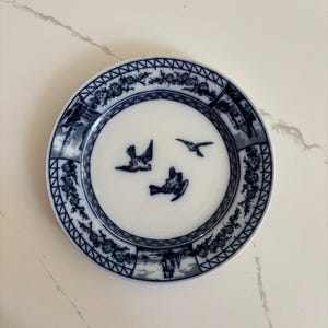 May include: A round ceramic plate featuring a white center with three dark blue birds in flight. The rim is decorated with a dark blue pattern of floral motifs, architectural structures, and a ship, all set against a white background.