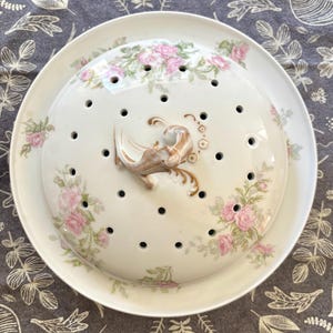 May include: A round white porcelain cheese dome with a matching plate, featuring a delicate pink rose floral pattern and green foliage. The dome has numerous small ventilation holes and a central decorative handle with gold accents.