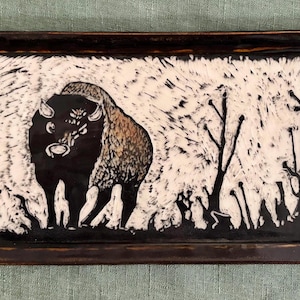 May include: A rectangular ceramic tray with a dark brown glazed rim and a central illustration of a black bison standing in a white field with black, stylized trees and brush. The artwork features high-contrast, textured black and white lines.