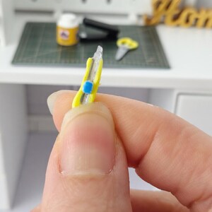 May include: A miniature yellow utility knife with a blue button is held in a hand. The knife has a silver blade and is set against a blurred background of a desk and other miniature craft supplies.