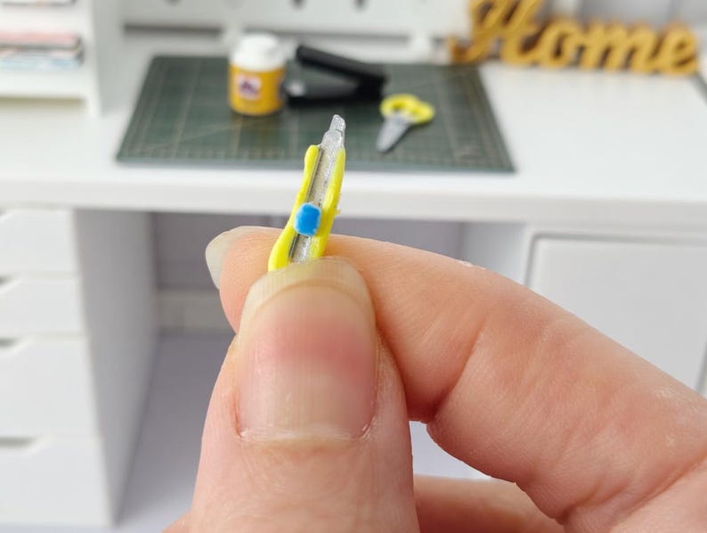 May include: A miniature yellow utility knife with a blue button is held in a hand. The knife has a silver blade and is set against a blurred background of a desk and other miniature craft supplies.