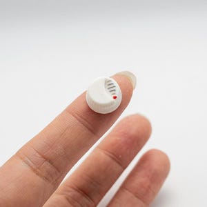 Könnte beinhalten: Ein kleines, weißes, kreisförmiges Objekt mit einer Reihe vertikaler Linien und einem roten Punkt. Das Objekt, das einem Miniatur-Rauchmelder ähnelt, wird zwischen zwei Fingern vor einem weißen Hintergrund gehalten. Es ist wahrscheinlich ein Miniaturmodell.