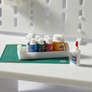 May include: Miniature craft supplies including small bottles of paint in various colors, a bottle of glue with a red tip, and a container of yellow paint. The items are arranged on a green cutting mat and a white tray.
